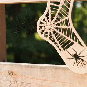 K&ouml;nnte beinhalten: Ein Holzausschnitt einer Socke mit einem Spinnennetz-Design und einer Spinne in der Mitte. Der Ausschnitt h&auml;ngt an einem Holzzaun.