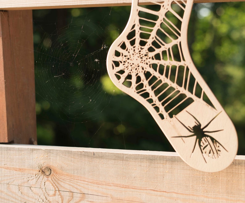 K&ouml;nnte beinhalten: Ein Holzausschnitt einer Socke mit einem Spinnennetz-Design und einer Spinne in der Mitte. Der Ausschnitt h&auml;ngt an einem Holzzaun.