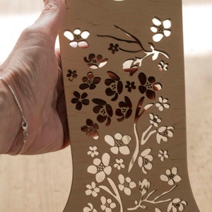 May include: A wooden cutout of a floral design with a delicate pattern of flowers and branches. The cutout is light brown and has a smooth finish.