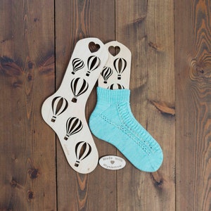 Può includere: Due blocca calze in legno a forma di cuore con ritagli di mongolfiere. Un calzino di lana blu chiaro è teso su uno dei blocca calze. Il blocca calze ha il testo "Made with MyKnitrow".