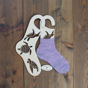 Op de afbeelding: Een paar paarse en witte gebreide sokken op een houten sokkenhanger met een schildpadontwerp. De hanger heeft de tekst "Made with MyKnitrow" erop.