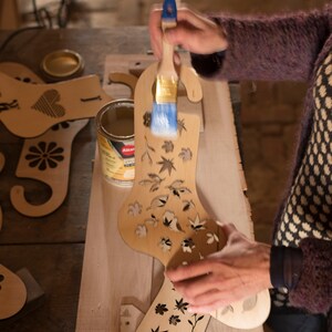 May include: A person is applying a clear sealant to a wooden Christmas stocking ornament with a brush. The stocking is decorated with a cut-out design of leaves and flowers.