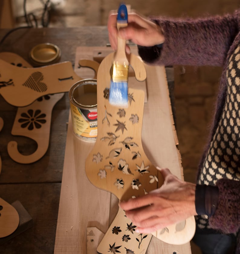 May include: A person is applying a clear sealant to a wooden Christmas stocking ornament with a brush. The stocking is decorated with a cut-out design of leaves and flowers.