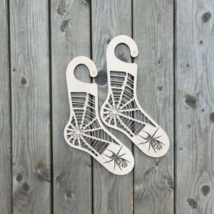 K&ouml;nnte beinhalten: Zwei h&ouml;lzerne Sockenhalter in Form von Socken mit einem Spinnennetz-Design und einer Spinne auf jedem. Die Halter sind mit "M" f&uuml;r mittlere Gr&ouml;&szlig;e gekennzeichnet.