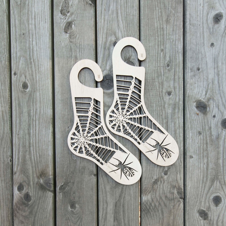 K&ouml;nnte beinhalten: Zwei h&ouml;lzerne Sockenhalter in Form von Socken mit einem Spinnennetz-Design und einer Spinne auf jedem. Die Halter sind mit "M" f&uuml;r mittlere Gr&ouml;&szlig;e gekennzeichnet.
