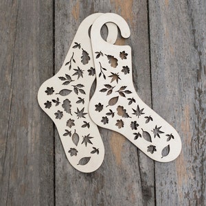 May include: Two wooden sock forms with a leaf pattern cut out. The forms are light brown and have a hanger at the top.