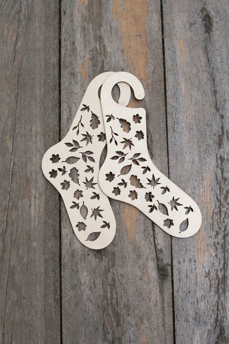 May include: Two wooden sock forms with a leaf pattern cut out. The forms are light brown and have a hanger at the top.