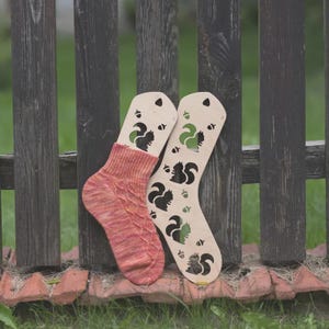 Può includere: Un paio di forme per calzini in legno con ritagli di scoiattoli si trova accanto a un paio di calzini a fantasia rossi e arancioni. Le forme in legno presentano disegni di scoiattoli neri e verdi. I calzini poggiano contro una staccionata di legno usurata.