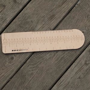 May include: A wooden ruler with a cm and inch scale, a row of holes for knitting needles, and the text "cm scale" and "inch scale".