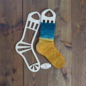 Op de afbeelding: Twee houten sokkenspanners met een paar handgebreide sokken in geel, blauw en groen. De sokken zitten op de spanner en een klein label met de tekst "Made with ♥ Mlynnjedrow".