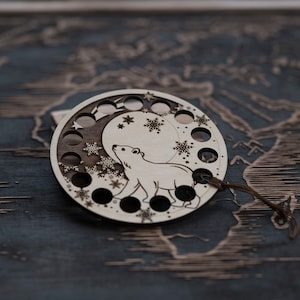 Może przedstawiać: Drewniana ozdoba przedstawiająca niedźwiedzia polarnego stojącego na rosnącym księżycu z płatkami śniegu i gwiazdami. Ozdoba ma 12 otworów wokół krawędzi.