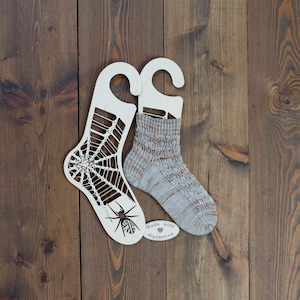 May include: Two wooden sock blockers, one with a spider web design and a spider, the other plain. A pair of knitted socks is displayed on the blockers. The socks are a light grey with brown stripes. A small wooden tag reads "Made with love Mlynjadrow".