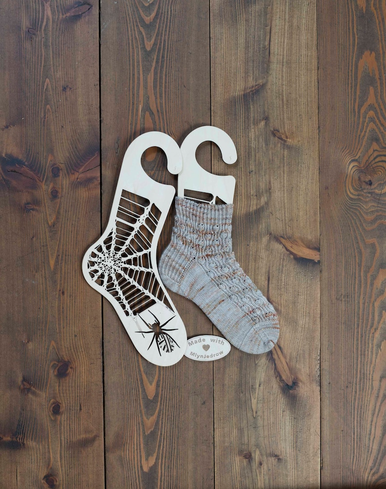 K&ouml;nnte beinhalten: Zwei h&ouml;lzerne Sockenspanner, einer mit einem Spinnennetz-Design und einer Spinne, der andere schlicht. Ein Paar gestrickte Socken wird auf den Spannern pr&auml;sentiert. Die Socken sind hellgrau mit braunen Streifen. Ein kleines Holzschild tr&auml;gt die Aufschrift "Made with love Mlynjadrow".