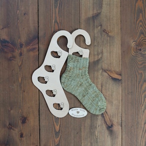 Op de afbeelding: Twee houten sokkenhangers met een groene en bruine sok op een van de hangers. De hangers hebben een schapenontwerp en de tekst "Made with ♥ Mlynnjadrow" staat op een klein ovaal etiket.