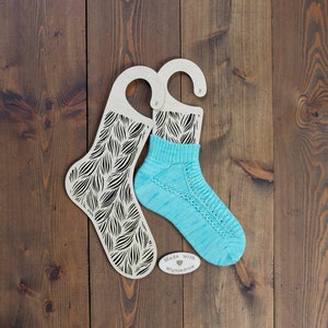 Può includere: Due bloccanti per calze in legno con un motivo a foglie nere, uno con un calzino di lana blu chiaro all'interno. I bloccanti per calze hanno un gancio in alto e il testo "Made with ♥ Mlynnjedrow" è su un piccolo cartellino di legno.