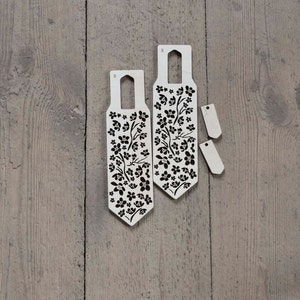 May include: Two wooden tags with a floral design and the letter "M" printed on them. The tags are on a wooden surface.