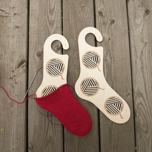 May include: Two wooden sock drying forms with a red knitted sock on one of the forms. The forms have a cut-out design of yarn balls.