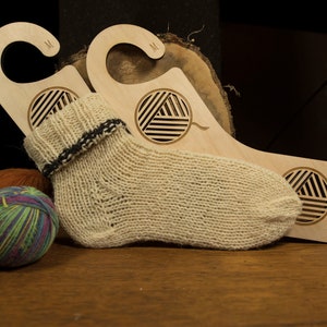 May include: Two wooden sock drying hangers with a yarn ball design. The hangers are labelled "M" and have a hook at the top. A beige knitted sock is draped over the hangers.