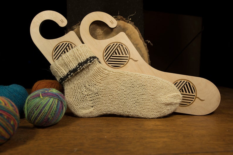 May include: Two wooden sock drying hangers with a yarn ball design. The hangers are labelled "M" and have a hook at the top. A beige knitted sock is draped over the hangers.