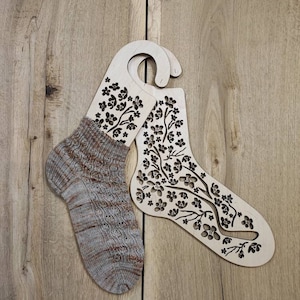May include: Two wooden sock blockers with a floral design. The blockers are shaped like a sock and have a hook at the top for hanging. One sock blocker is holding a knitted sock with a brown and white pattern.