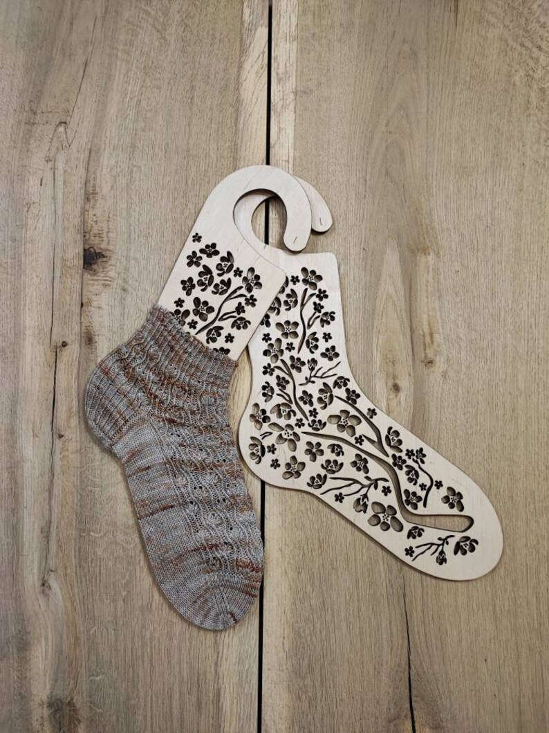 May include: Two wooden sock blockers with a floral design. The blockers are shaped like a sock and have a hook at the top for hanging. One sock blocker is holding a knitted sock with a brown and white pattern.