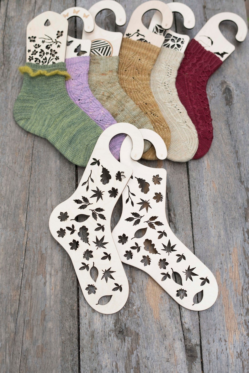 May include: Six pairs of knitted socks displayed on wooden sock blockers with leaf and floral designs. The socks are in various colors, including green, purple, brown, beige, and red.