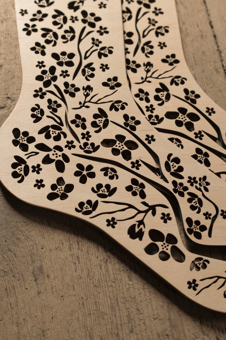 May include: A pair of wooden sock-shaped ornaments with a delicate floral design cut out. The design features small black flowers and branches on a light brown background.
