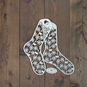 Op de afbeelding: Twee witte metalen sokken-uitsnijdingen met een bloemenpatroon. De uitsnijdingen bevinden zich op een bruin houten oppervlak. De tekst "Made with ♥ Mlynjedrow" bevindt zich onderaan de uitsnijdingen.