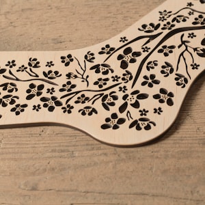 May include: A wooden sock block with a floral design. The block is made of light wood and has a black floral pattern cut out of it. The block is designed to help knitters shape socks.