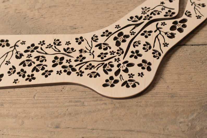 May include: A wooden sock block with a floral design. The block is made of light wood and has a black floral pattern cut out of it. The block is designed to help knitters shape socks.
