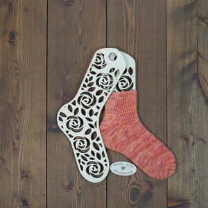 May include: A pair of orange and pink hand-knitted socks on a wooden sock blocker with a white label that says "Made with ♥ Mlynjedrow".