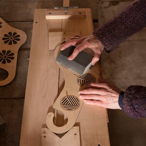 May include: A person sanding a wooden cutout of a stocking with a sanding block. The stocking has a geometric design and a hook at the top.