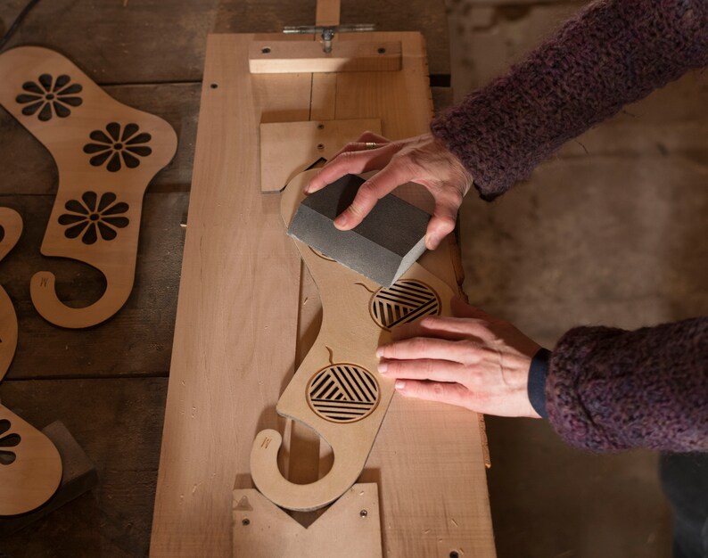 May include: A person sanding a wooden cutout of a stocking with a sanding block. The stocking has a geometric design and a hook at the top.