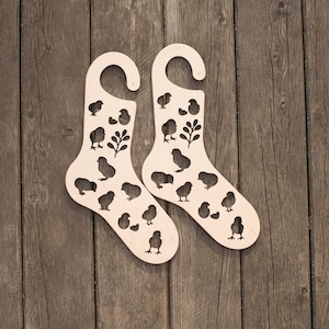 Può includere: Un paio di appendini per calze in legno con un design a forma di pulcini e uova ritagliato. Gli appendini sono di colore marrone chiaro e hanno un gancio nella parte superiore.