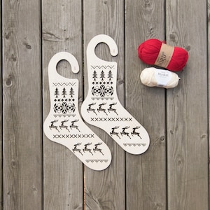 May include: Two white wooden Christmas stocking ornaments with a reindeer and snowflake design. The ornaments have a hook for hanging. Two balls of yarn are in the background, one red and one white.