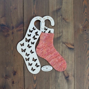 Può includere: Due portacalze in legno con tagli a forma di farfalla, uno con un paio di calze lavorate a mano rosa e arancioni. I portacalze hanno il testo "Made with Miynjedrow" su di loro.