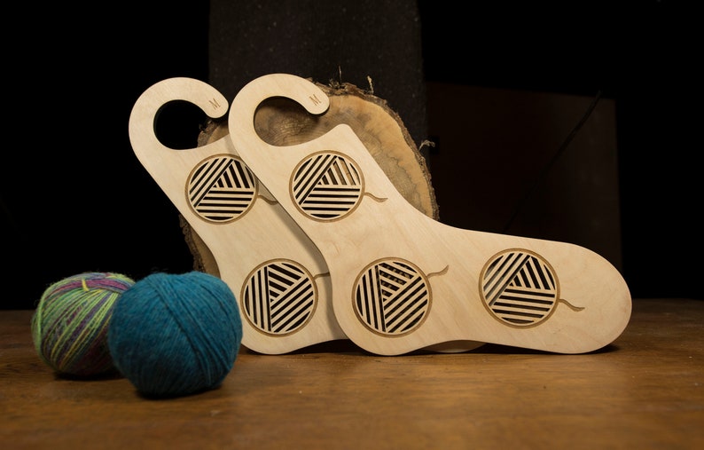 May include: Two wooden sock holders with a yarn ball design. The holders are light brown and have a hook at the top. The holders are sitting on a wooden surface with two balls of yarn.