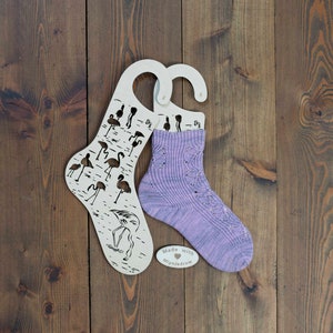 May include: A pair of purple and white knitted socks on a wooden sock hanger with a flamingo design. The hanger has the text "Made with MyNeedle" on it.