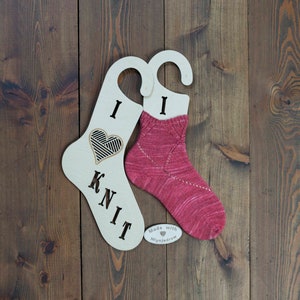 Può includere: Due appendini per calze in legno con le parole "I Knit" e un design a forma di cuore. Un appendino ha una calza rosa appesa. Gli appendini sono su uno sfondo di legno. La calza è realizzata con filato Miynjadrow.