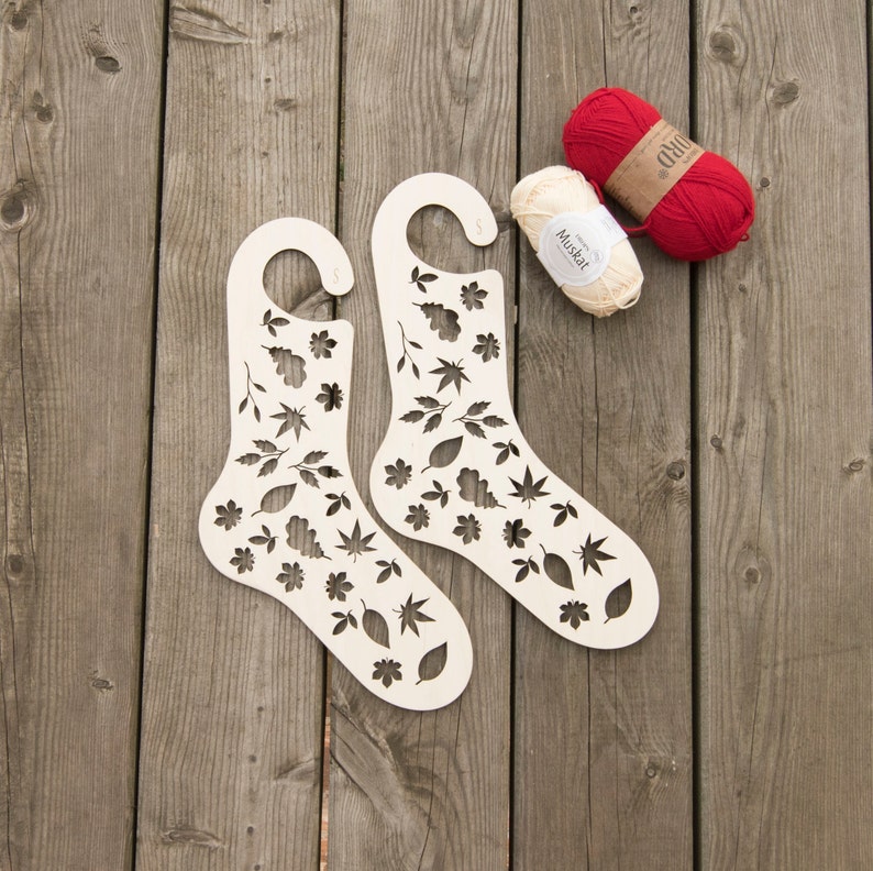 May include: Two wooden sock blockers with a leaf and flower design. The blockers are on a wooden surface with two balls of yarn, one red and one white.