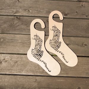 Op de afbeelding: Twee houten schoenhouders in de vorm van een tijger, met een ophanglus aan de bovenkant. De houders zijn gemaakt van lichtbruin hout en hebben een zwart-wit tijgerontwerp.