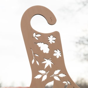 May include: A brown wooden hanger with a cut-out design of leaves and flowers. The hanger has a hook at the top and the letter "S" is visible on the top right corner.