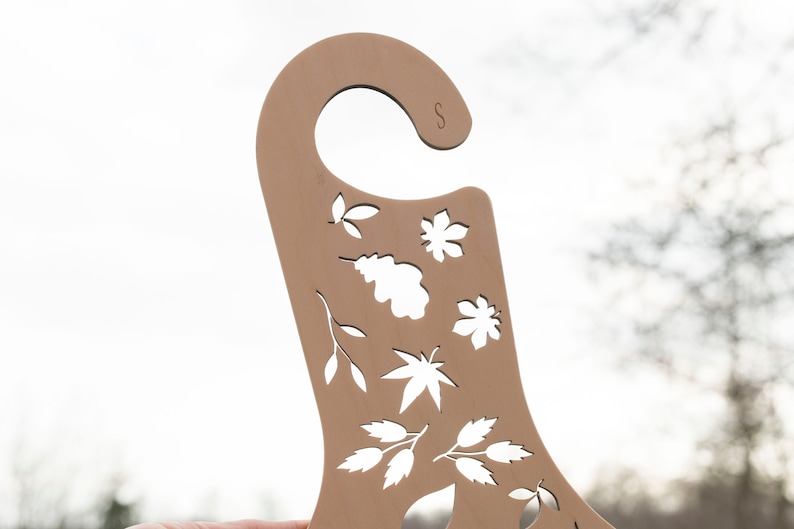 May include: A brown wooden hanger with a cut-out design of leaves and flowers. The hanger has a hook at the top and the letter "S" is visible on the top right corner.
