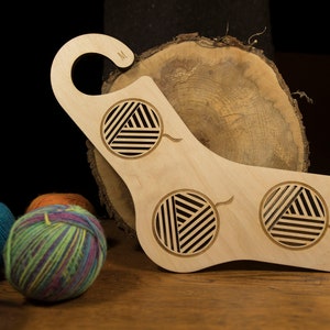 May include: A wooden yarn bowl with three cut-out circles for yarn balls. The bowl is light brown and has a hook on the top. The bowl is sitting on a wooden surface with three balls of yarn in the foreground.