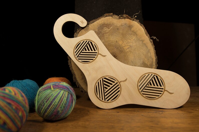 May include: A wooden yarn bowl with three cut-out circles for yarn balls. The bowl is light brown and has a hook on the top. The bowl is sitting on a wooden surface with three balls of yarn in the foreground.