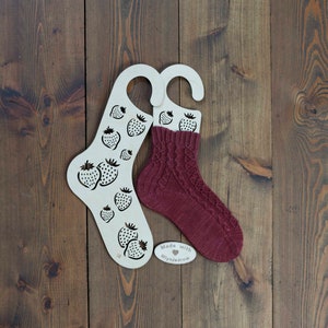 Op de afbeelding: Twee houten sokkenspanners in de vorm van aardbeien met een paar bordeauxrode gebreide sokken op een van de spanner. Een klein houten label met de tekst "Made with love Mlynjadrow" ligt op de tafel.