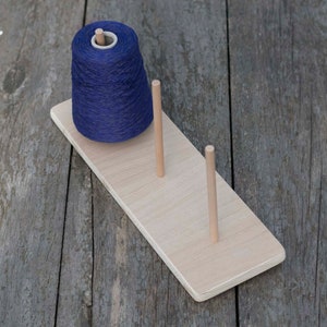 Puede incluir: Un soporte de hilo de madera con tres clavijas y una bola de hilo azul.