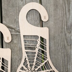 K&ouml;nnte beinhalten: Zwei Sockenhalter aus Holz mit einem Spinnennetz-Design. Jeder Halter hat einen Haken oben und der Buchstabe "M" ist in den Haken graviert.