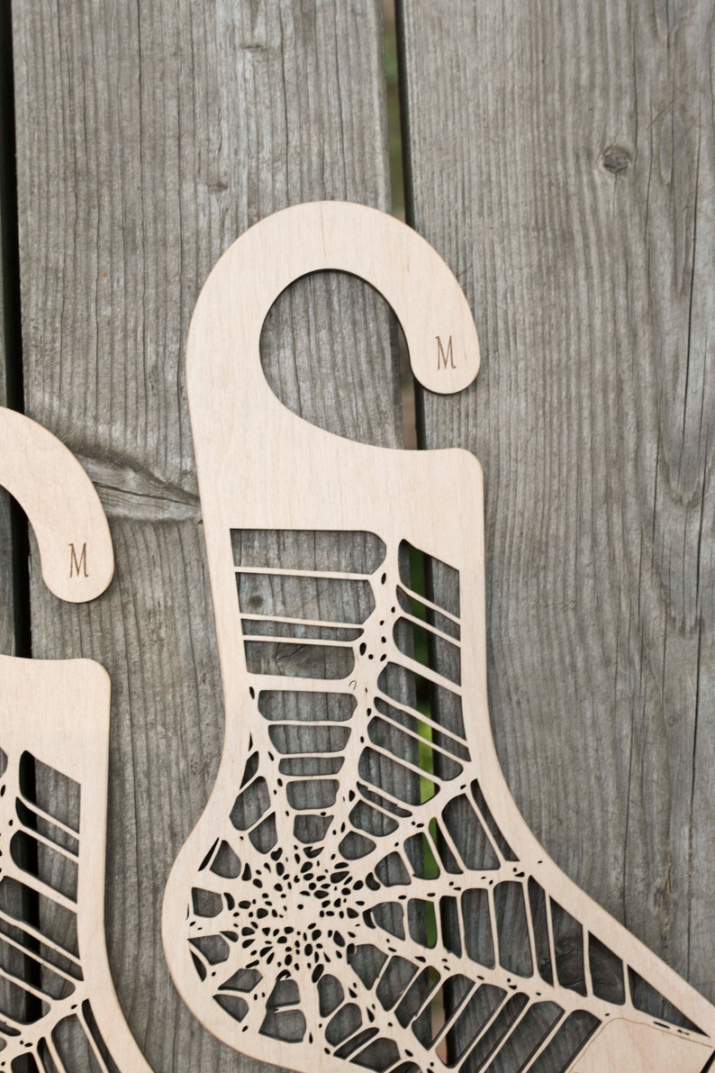 K&ouml;nnte beinhalten: Zwei Sockenhalter aus Holz mit einem Spinnennetz-Design. Jeder Halter hat einen Haken oben und der Buchstabe "M" ist in den Haken graviert.