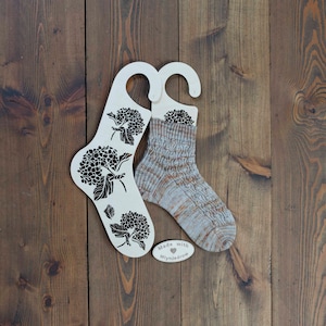 Op de afbeelding: Twee witte houten sokkenblokkers met een bloemenmotief en een klein label met de tekst "Made with ♥ Mlynjadrow".